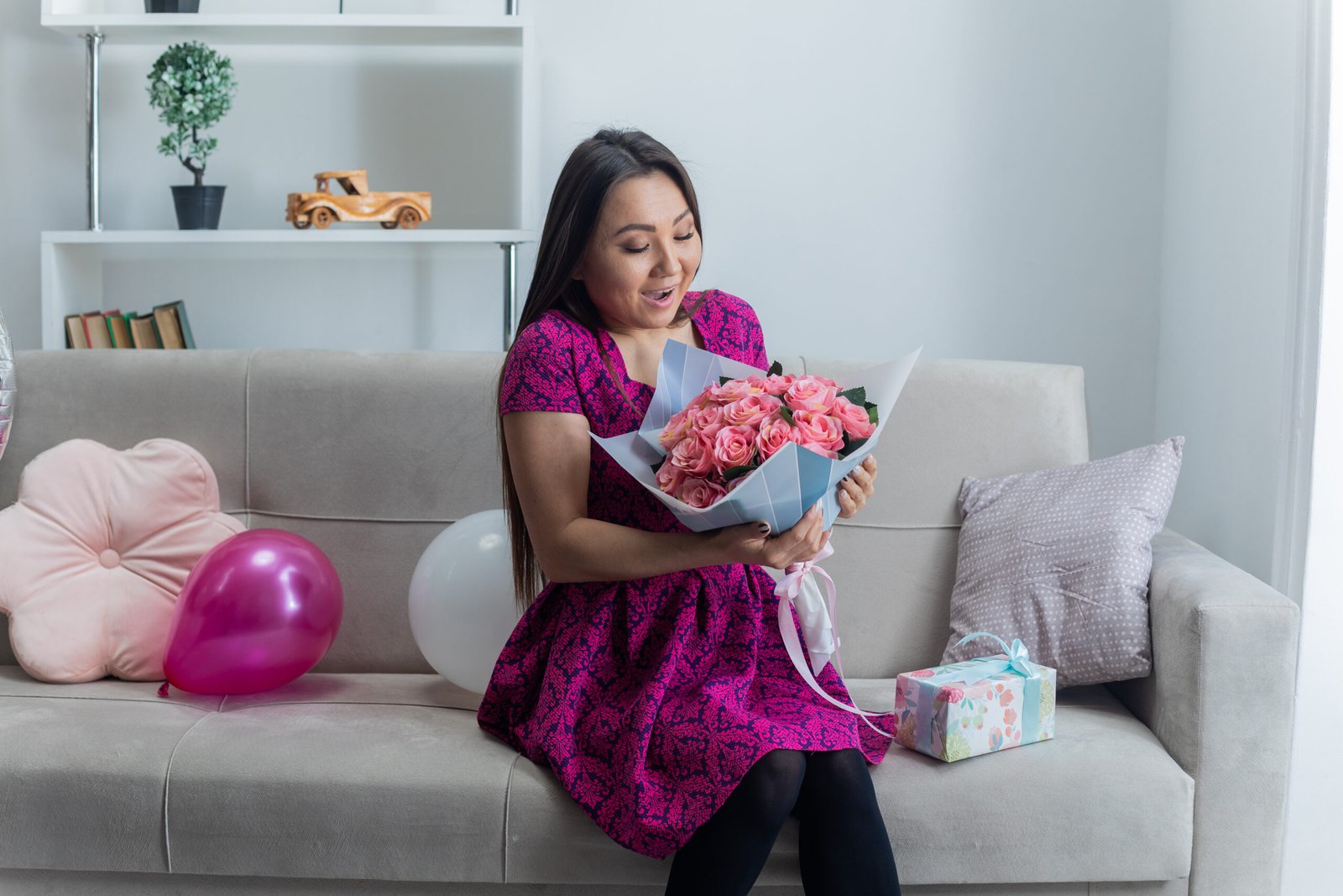 happy and excited asian young woman in beautiful dress smiling cheerfully sitting on a couch with bouquet of flowers and present in light living room celebrating international women's day march